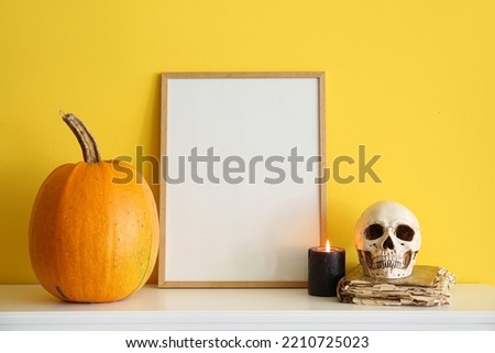 Blank frame with Halloween pumpkin, candle, skull and book on mantelpiece near yellow wall