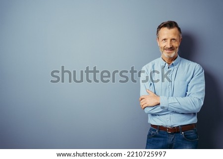 Handsome middle-aged man, standing with his arms folded, looking at camera and smiling. Half-length front portrait against blue background with copy space Royalty-Free Stock Photo #2210725997