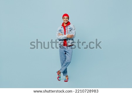 Full body young smiling middle eastern man 20s he wear denim jacket red hat hold hands crossed folded look camera isolated on plain pastel light blue cyan background studio. People lifestyle concept