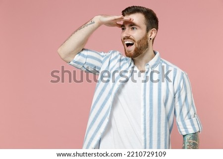Young smiling happy caucasian unshaven man 20s wear blue striped shirt white t-shirt hold hand at forehead look far away distance isolated on pastel pink color background studio. Tattoo translate fun.
