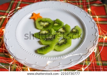Edible Christmas tree made of kiwi and carrots on a red tablecloth. Lights are burning around, a Christmas garland. High quality photo