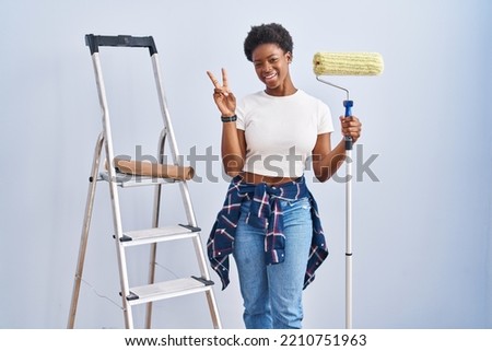 African american woman holding roller painter smiling with happy face winking at the camera doing victory sign with fingers. number two. 