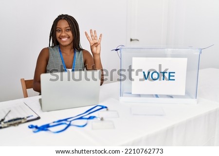 Young african american woman working at political election sitting by ballot showing and pointing up with fingers number four while smiling confident and happy. 