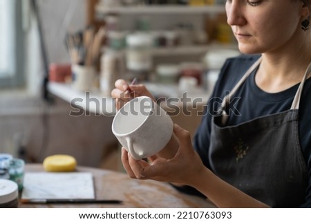 Craftswoman creates new masterpiece in work studio making sketch of future pattern on white mug. Lady master wants to decorate kitchen with painted clay dish using pencil to draw interesting pattern