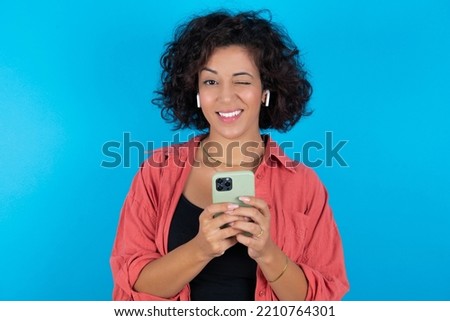 Pleased young beautiful woman with curly short hair wearing red overshirt over blue wall using self phone and looking and winking at the camera. Flirt and coquettish concept.
