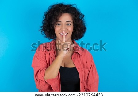 Smiling young beautiful woman with curly short hair wearing red overshirt over blue wall makes shush gesture, holds fore finger over lips hides secret. Be mute, please.