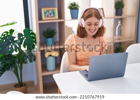 Young redhead woman using laptop and headphones sitting on table at home