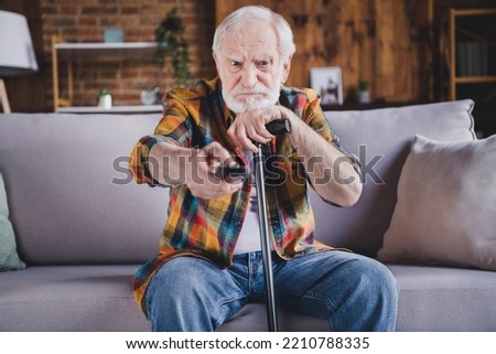 Photo of angry irritated man pensioner wear checkered shirt changing tv channels holding walking cane sitting sofa indoors house room