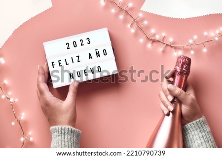 Text Happy New Year 2023 in Spanish Language on lightbox. Light garland, woman hands with lightbox and metallic pink champagne bottle. New Year, Sylvester celebration. Creative concept image, pink