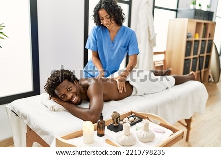 Young physiotherapist woman smiling happy giving back massage to african american man at the clinic. Royalty-Free Stock Photo #2210798235
