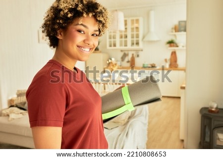 Side view of happy african american woman with afro hair smiling at camera, standing in her living-room with gray rolled mat in hands, preparing to practice yoga, pilates or fitness workout