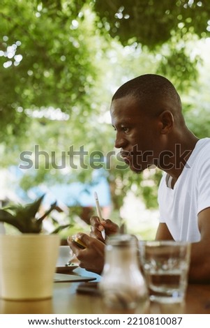 Vertical photo of concentrated modern dark skin man in casual clothes writing business plan in textbook during webinar on laptop in outdoor coffee shop. Businessman sitting with smartphone and PC