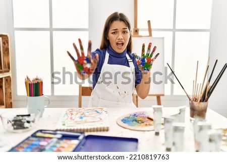 Young brunette woman at art studio with painted hands afraid and terrified with fear expression stop gesture with hands, shouting in shock. panic concept. 