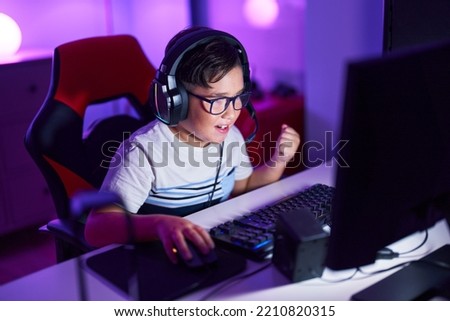 Adorable hispanic boy streamer playing video game with winner expression at gaming room