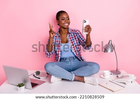 Full size portrait of cheerful nice girl sit desktop hold telephone make selfie show v-sign isolated on pink color background