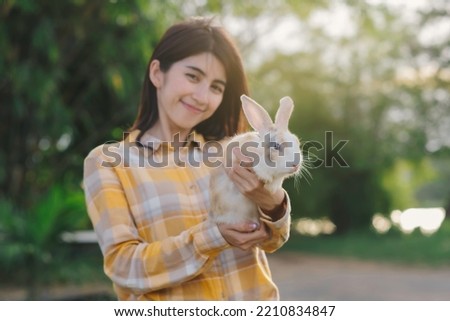 Close up cute and fluffy rabbit in woman's hand with backlit and blur asian woman in yellow scotish shirt looking at camera with smile and bokeh, orange light of sunset.