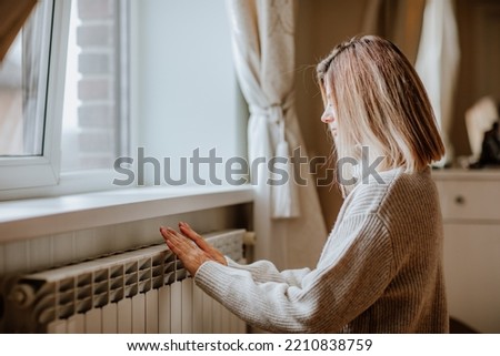 Young blond woman in long winter beige sweater is posing at home near the radiator. Winter season concept. Economy program  Royalty-Free Stock Photo #2210838759