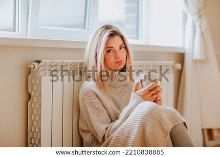 Young blond woman in long winter beige sweater is holding a cup of coffee and posing at home near the radiator. Winter season concept. Economy program and mental health concept Royalty-Free Stock Photo #2210838885