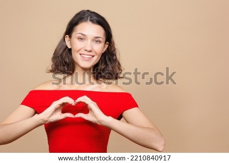 woman on a beige background smiles and makes a heart symbol with her hands. a happy person rejoices. a young European woman for a sale poster, a place for text. closed posture, resentment and