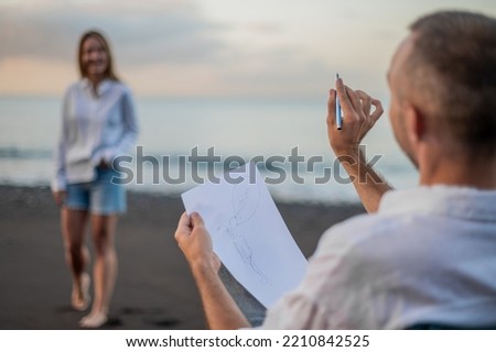 Young couple on a beach at sunset, she poses and he draws a picture of her