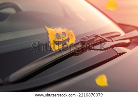 Cheerful yellow leaf with emotion, on the windshield of the car under the brush. Concept of warm autumn or Halloween holiday. Toned background Royalty-Free Stock Photo #2210870079