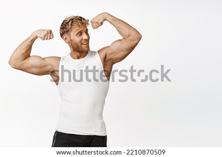 Handsome athlete looking strong, flexing biceps, showing his strong muscles arms, smiling pleased, workout and doing fitness exercises, white background Royalty-Free Stock Photo #2210870509