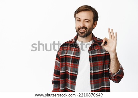 Handsome smiling man winks and shows okay sign, approves, likes and recommends smth good, praise promo deal, white background