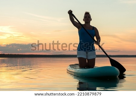 A woman on a kneeling SUP board with an oar at sunset against a golden sky floats in the water of the lake. horizontal photo
