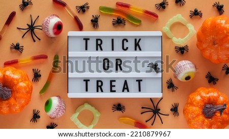 Pumpkins, cauldrons, pots, worms, jaws, spiders, eye-shaped candies and a text board with the inscription TRICK OR TREAT on a orange background, top view. A collection of items for a Halloween party