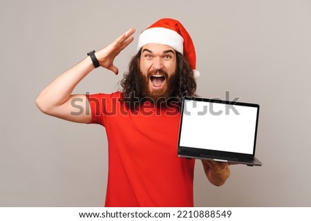Shocked man is showing a Christmas offer on a laptop screen he is holding. Studio shot over grey background.