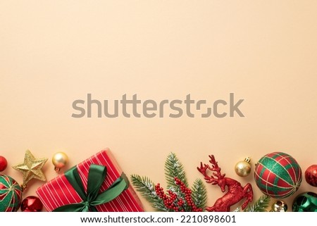 Christmas Eve concept. Top view photo of red giftbox with green ribbon bow baubles gold star reindeer ornament mistletoe berries and pine branch in frost on isolated beige background with empty space