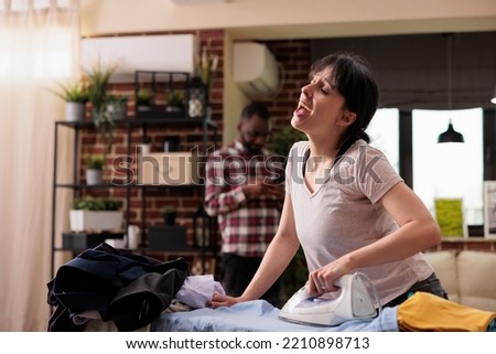Happy smiling woman singing and ironing clothes on board, husband on phone in background. Cultural diverse couple at home, cheerful housewife doing daily chores with positive vibes and emotions Royalty-Free Stock Photo #2210898713