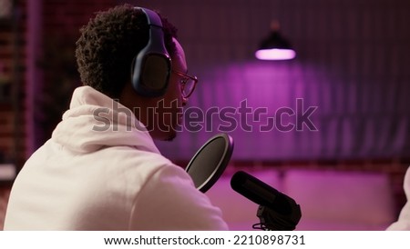 Over shoulder view of african american podcaster talking confident while broadcasting live with microphone. Static tripod shot of online radio host during interview listening and smiling in studio.