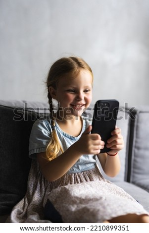 Portrait of cute little cute girl holding smartphone and resting at sofa at home. Choosing favorite music or cartoons,texting messages,browsing internet,watching video, playing games on mobile phone