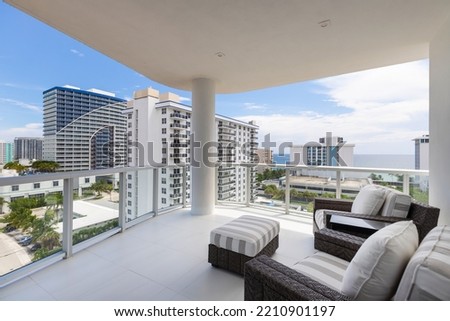 View of elegant balcony with wooden and wicker furniture in opaque colors, glass railing, with urban landscape and sea in the background, Royalty-Free Stock Photo #2210901197