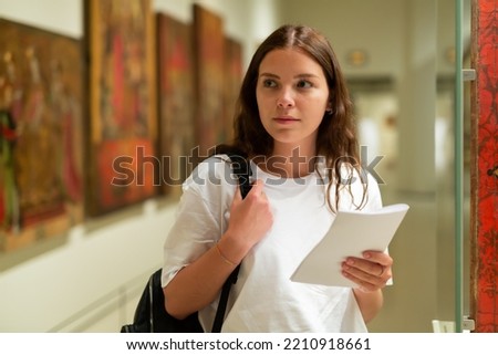 Portrait of a focused girl standing in the museum hall next to an exhibit located in a glass cabinet, with a booklet of the ..exhibition program Royalty-Free Stock Photo #2210918661