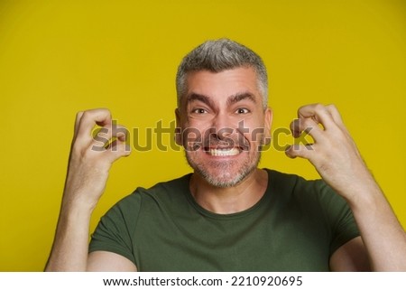 Middle aged caucasian grey haired man showing anger facial expression with hands up wearing khaki t-shirt isolated on yellow background. Handsome man showing his grin with perfect teeth. Royalty-Free Stock Photo #2210920695
