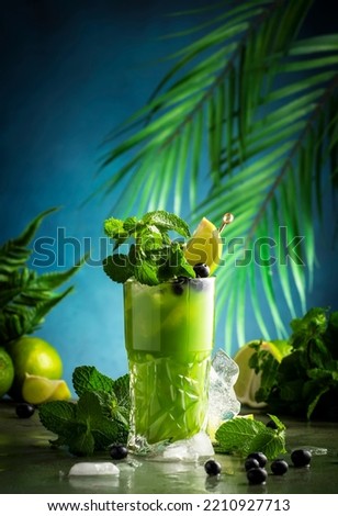 Green alcoholic cocktail with gin, tequila, liquor, juice, lime, blueberry, mint and ice. Dark tropical background, copy space