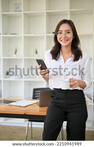 Portrait, Gorgeous and successful millennial Asian businesswoman or female manager standing in her office, holding her smartphone and a coffee cup.