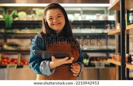 Shop employee with Down syndrome smiling at the camera while holding a digital tablet. Happy woman with an intellectual disability working as a storekeeper in a local grocery store. Royalty-Free Stock Photo #2210942953