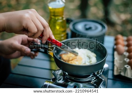 Portrait of Asian traveler man glasses frying a tasty fried egg in a hot pan at the campsite. Outdoor cooking, traveling, camping, lifestyle concept. Royalty-Free Stock Photo #2210964541
