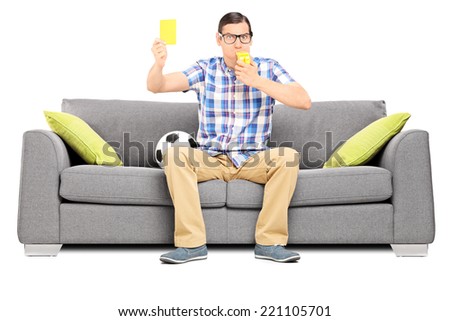 Angry football fan holding a yellow card seated on couch isolated on white background