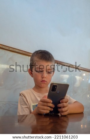 little boy holds the mobile phone with both hands and stares at it, because he is amused by it. behind him there is a wall of white tiles.