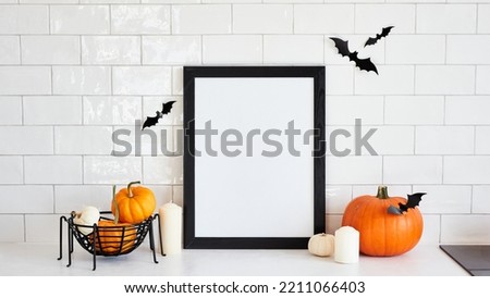 Vertical black picture frame mockup with pumpkins and halloween home decor on white desk table in scandinavian kitchen interior. Halloween holiday poster design.