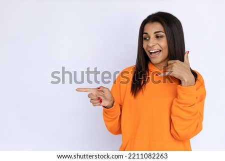 Portrait of happy Hispanic woman making call me gesture. Cheerful young model with long hair in orange hoodie looking away, smiling and pointing finger. Advertisement concept.