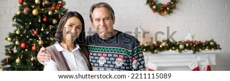 senior man hugging multiracial wife while smiling at camera near decorated christmas tree, banner