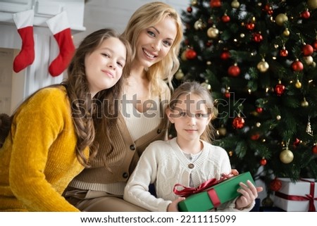 happy girls with blonde mother looking at camera near decorated christmas tree on blurred background