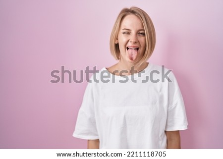 Young caucasian woman standing over pink background sticking tongue out happy with funny expression. emotion concept. 