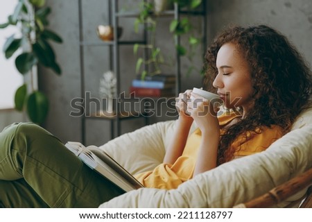 Sideways young woman of African American ethnicity wear t-shirt drink coffee read book close eyes sits in armchair stay at home flat rest relax spend free spare time in living room indoor grey wall Royalty-Free Stock Photo #2211128797