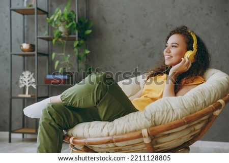 Side view dreamful minded young happy woman of African American ethnicity wear t-shirt headphones listen to music sits in armchair stay at home rest relax spend free spare time in living room indoors